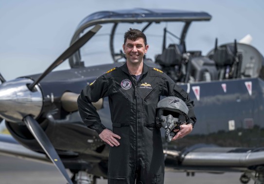 A military pilot smiles broadly whilst wearing a black flying suit. He stands in front of a small black aircraft with one hand on his hip and the other holding his helmet. 