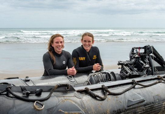 S1 Katrina Koch-Underhill (left) and S1 Sarah Gunderson (right) leaning on a Zodiac in front of the waves at Piha Beach.