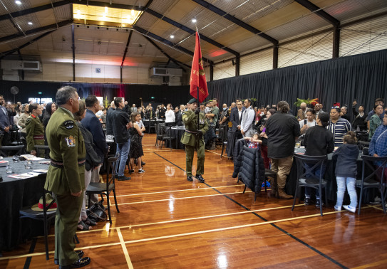 A soldier carrying a red flag walks down the centre of a hall as people stand next to the tables they were sitting at.