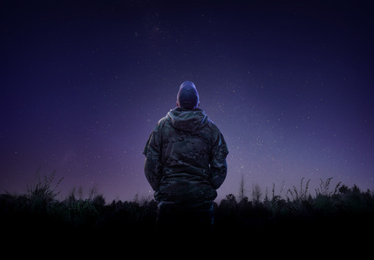 An aviator from the Royal New Zealand Air Force looks to the stars while wearing a camouflage jacket and a cap that has the letters 'RNZAF' on the back. 