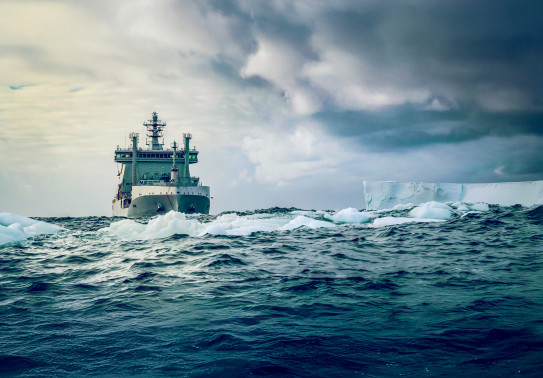 HMNZS Aotearoa seen bow on in grey green Antarctic seas which meet grey and cloudy skies about halfway. Light sea ice floats ahead of the ship and a distant flat topped, square angled ice shelf projects into the scene from the right.