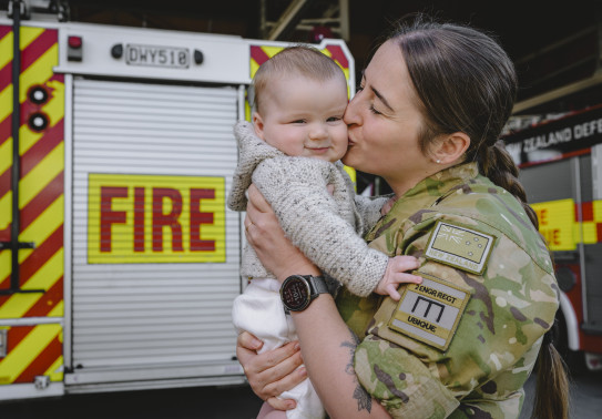 A soldier in uniform with eyes closed kisses baby who's looking at the camera. There's a fire truck in the background which says 'fire' on it. 