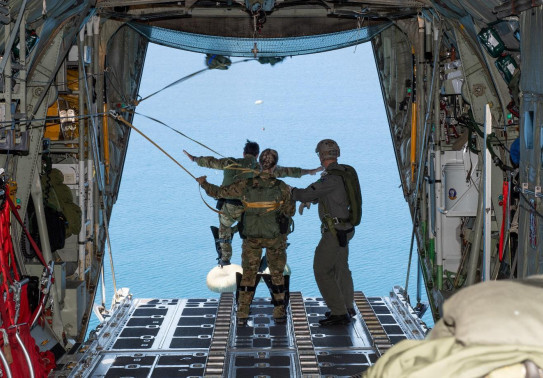 Uniformed people stand on the edge of an aircraft door ready to jump out over water. One person has already started to jump.  