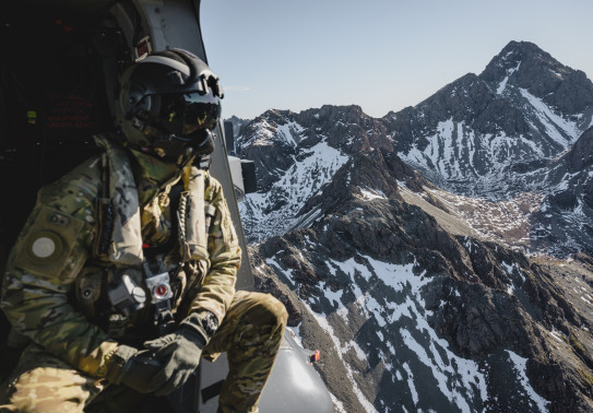 A helicopter loadmaster in full Air Force uniform including a flight helmet looks out the side of the aircraft to snow-capped mountains.