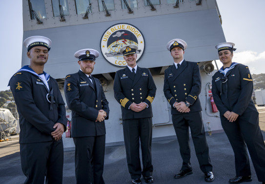 Five Navy sailors stand in uniform in front of a large "USS Blue Ridge". The ship casts a shadow on the sailors on a cold sunny day.