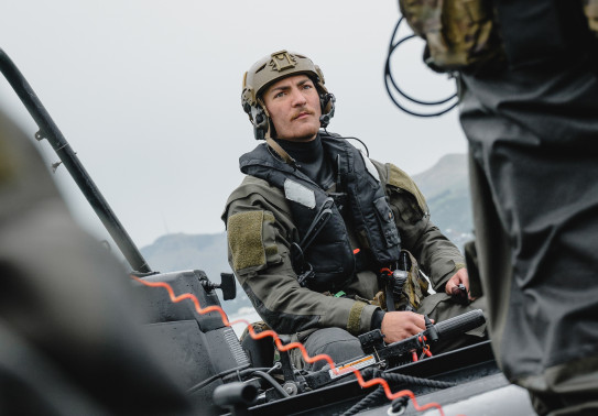 A soldier wearing full wet weather Army uniform including a life jacket and helmet is photographed between two other soldiers as they sit in small boats under grey skies.