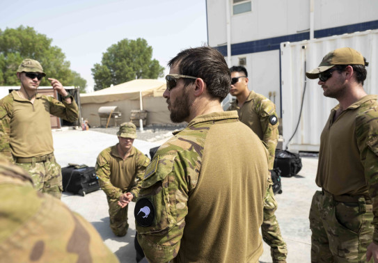 Soldiers stand in a circle for a brief, one of the soldiers is knelt down, containers, tents a prefabricated buildings surround the team under harsh sunlight.