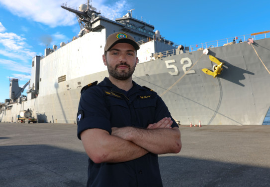 LT Cunningham stands with arms crossed in front of the large grey US Navy ship, USS Pearl Harbor. The number 52 is painted on the front-side of the ship.