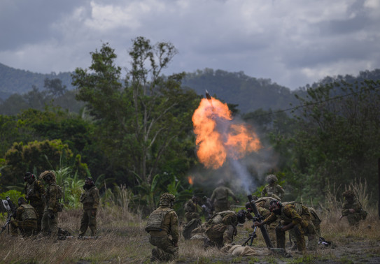 Army crews fire a mortar in a field in front of trees. The soldiers firing, are crouched down as a large flame and a round is launched from the mortar.