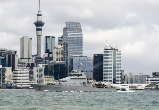 A grey ship sails in front of the Auckland city skyline.