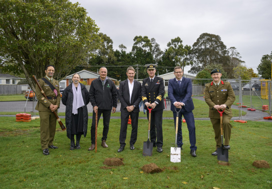 A line of seven people standing on grass, the four to the right are men holding spades. Several sods have been turned and are sitting on the grass in front of the people.