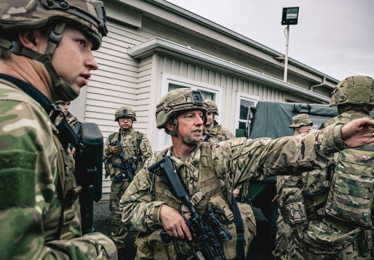Soldiers are in between vehicles out the front of a white panelled single-story building, one soldier points to the right while others listen.