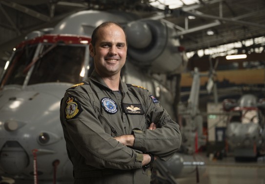 Male wearing an aircrew uniform smiles as he stands with his arms folded in front of a grey military helicopter. 