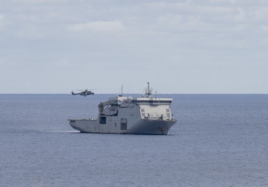 A large grey naval ship moves through the waters as a helicopter hovers above.