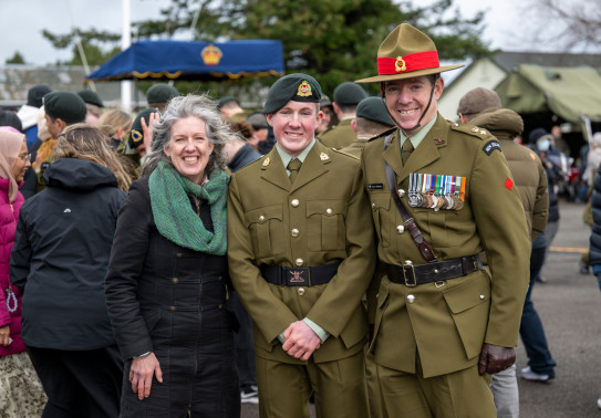  Proud Mum in a jacket and green scarf on the left, while the son and Dad (on the right) are in Army uniforms on the parade ground in front of other families and soldiers.