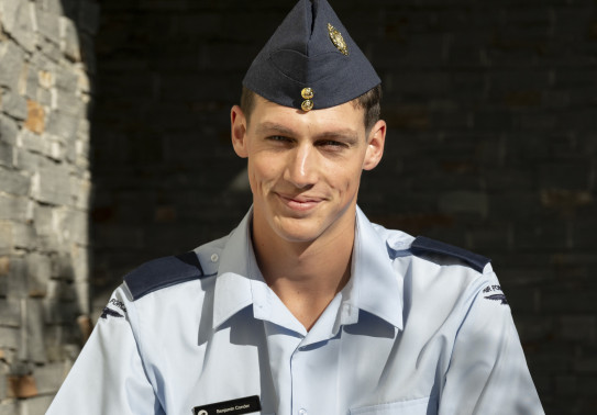 An aviator looks directly at the camera with a smile wearing his RNZAF uniform. In the background you can see a brick wall with sunlight on it and then a shadow directly behind the person.  