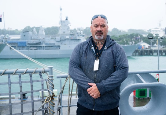A male stands with his arms clasped in front of him, he is wearing a casual grey hoodie and jeans and a lanyard with his sunglasses on his head. He stands aboard a military naval ship and behind him is another grey ship alongside the wharf with glimpses o