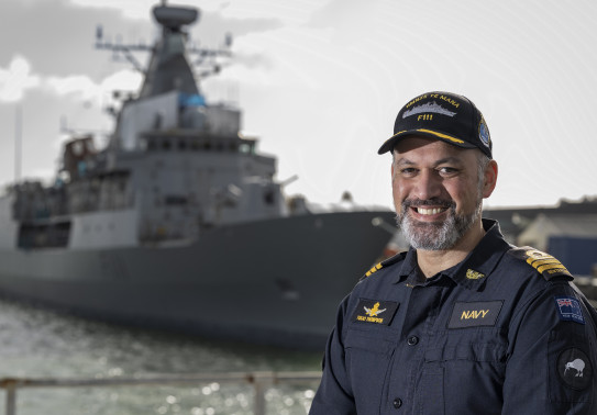 A uniformed sailor stands looking at the camera with a Navy ship in the background, out of focus. The sailor has a cap on and is smiling at the camera. 