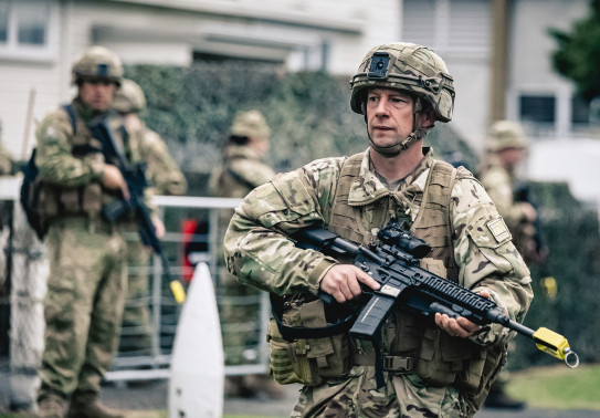 A soldier in full Army uniform and holding a weapon is in focus, while blurred in the background are more soldiers and a large bush in front of a building.