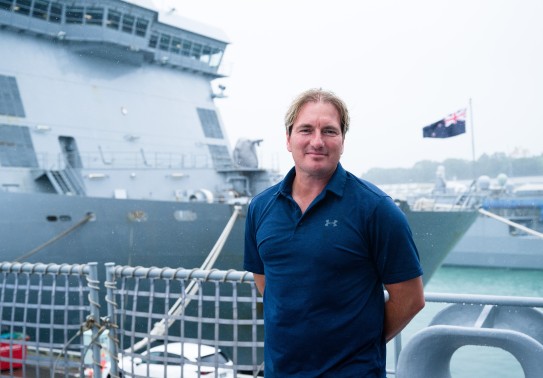 A blonde haired, middle aged man stands in front of two large grey naval ships.  He wears a navy blue under armour polo shirt and stands casually with his arms behind his back. Behind him the New Zealand flag flies prominently on the bow of the closer shi
