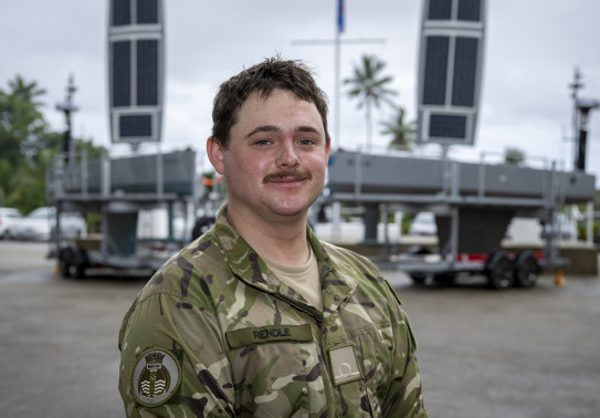 A man with a mostache in military uniform looks at the camera as he stands in front of the two grey USVs on trailers.