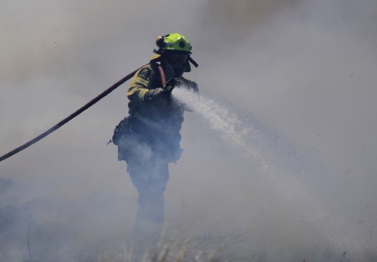 A firefighter emerges from the smoke with their hose on as it continues to spray water. 