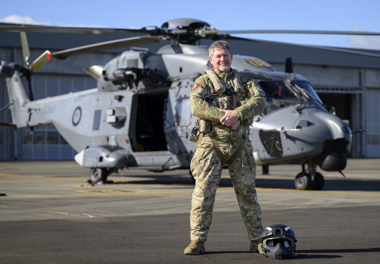 A uniformed aviator stands in front of a NH90 helicopter, looking and smiling at the camera, hands together. 