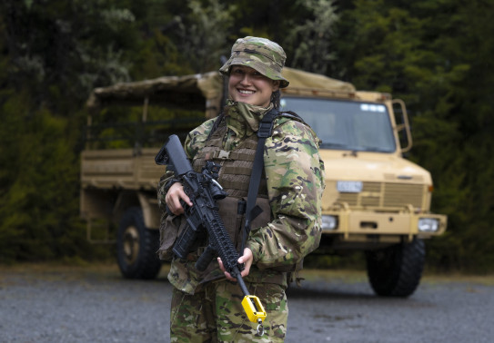 Flying Officer Celine Conaglen stands holding a MARS-L in camouflage uniform in front of a tan-coloured military truck.