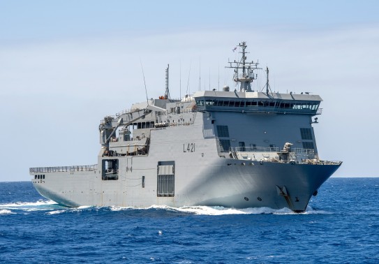 A large grey ship sails through blue water under clear skies,