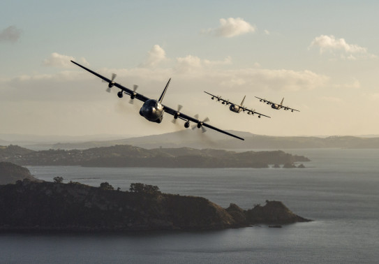 Three C-130H aircraft flying formation in a line over a harbour. All three aircraft are banking in a turn.
