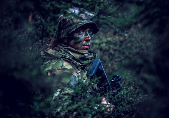 Female soldier standing camouflaged in bush.