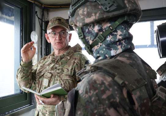 Two soldiers in different military uniform talk in a small room with large windows.
