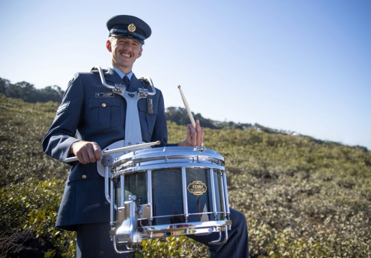 Percussionist Corporal Jeremy Richardson stands with a drum, all in uniform with greenery and sky in the background. 
