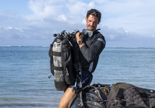 A diver holds a dive tank while in a dive t-shirt and shorts with a pair of goggles around his neck.