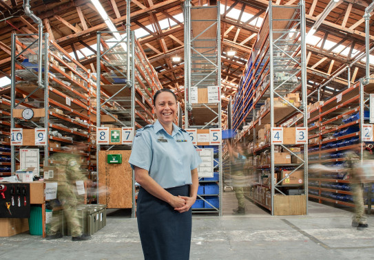 A woman stands in a warehouse with her hands together in front of her, she is wearing a RNZAF uniform. In the background, people in uniform are blurred by motion as industrial shelving, nets, trays and boxes. are lit by skylights in the ceiling.