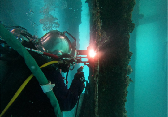 A diver undertakes a welding task in turquoise water. The diver is in a diving helmet and full diving and safety apparatus.