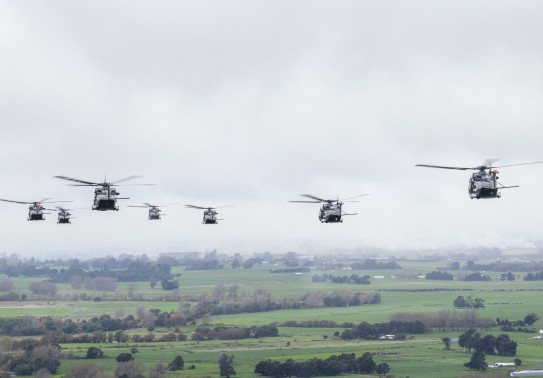 Seven of the Royal New Zealand Air Force’s NH90 helicopters fly in the sky over green land. The sky is very cloudy and grey. The image of the aircraft is taken from behind so the perspective is the back of al the aircraft. 