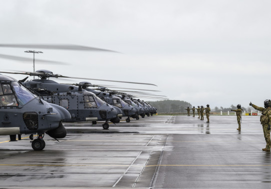 Nh90 helicopters line up on the flight line in cloudy weather. The ground is also wet. Aviators in uniform and helmets stand with thumbs up and arms out towards the aircraft. 