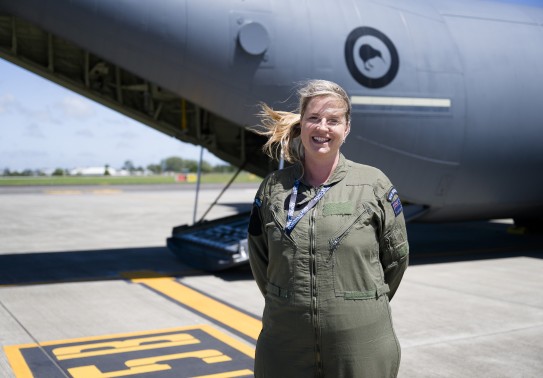 A woman stands on the tarmac smiling at the camera, her blond ponytail blowing in the wind while she wears a green flying suit with the New Zealand patch on her left arm.  A large grey aircraft is behind her, the ramp is down and the New Zealand Kiwi is o