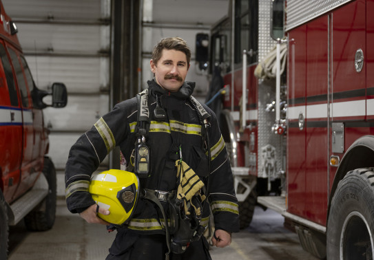 A firefighter stands in uniform, with a helmet in his hand, between two fire trucks in a fire station.