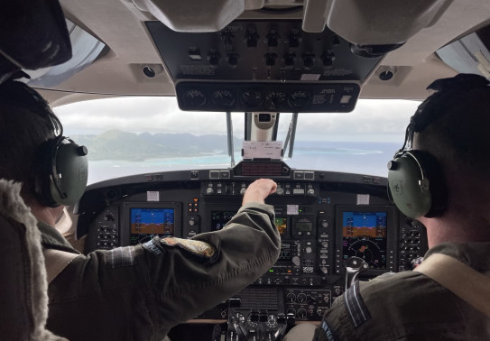 Two people in the flight deck of an aircraft, looking out the window. One of them is touching something on the panels. 