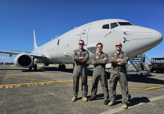 Three aviators in Air Force uniform stand with arms crossed in front of a large grey jet-powered aircraft under blue skies.