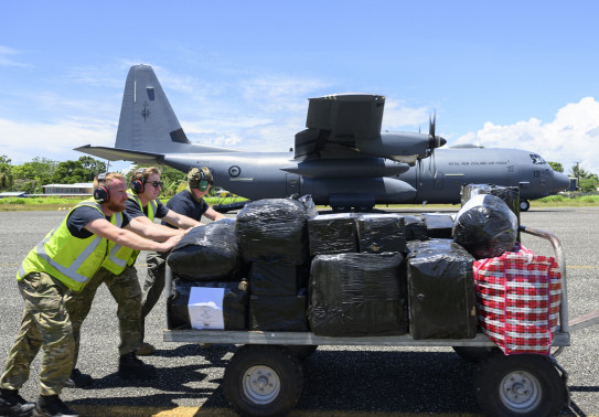Three people push a trolley full of cargo in front of a large grey aircraft.