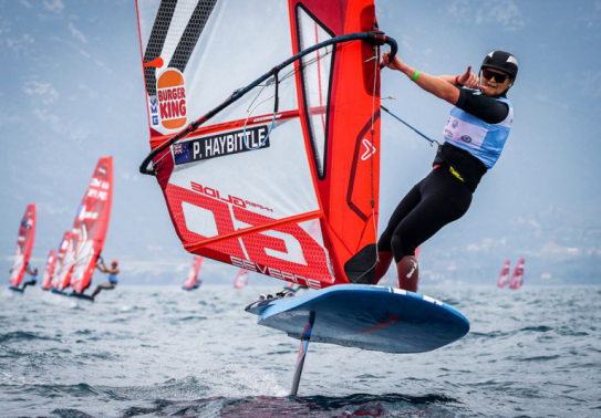 Corporal Haybittle on his windfoil, a blue board with a mat travelling down to the water and a red and white kite on top. He is pulling the shaka and is wearing a black helmet, glasses and wetsuit.