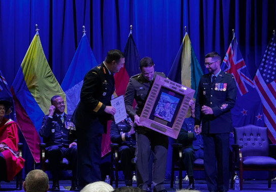 Major Prouse recieved a trophy on stage from two other military personnel. The lighting is very blue and flags line the back of the stage in front of a stage curtain.