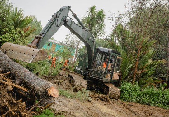A bulldozer sits atop mud as it pushes a pile of tree debris to the side. Several workers wearing high vis safety gear work in the background.