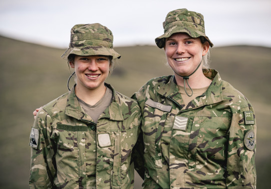 Two solders in uniform stand side-by-side, smiling at the camera. The background is blurred. 
