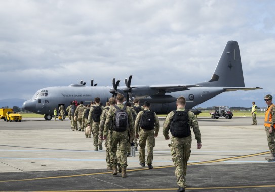 A group of soldiers walk toward a large grey military aircraft, they walk in two files, black backpacks on as military staff stand alongside with high vis vests.