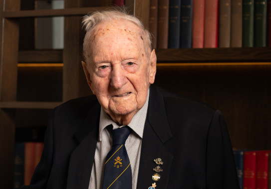 A man wearing a suit and medals looks at the camera as he stands in front of a bookshelf.