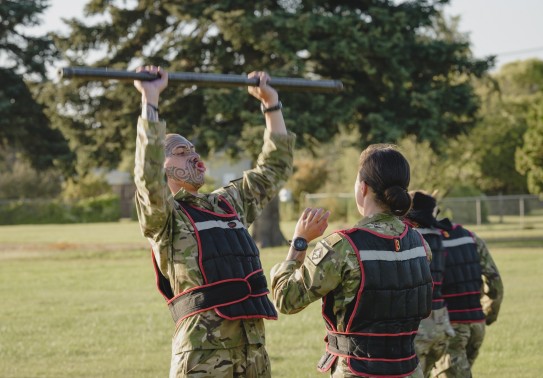 Two soldiers partner up completing a physical challenge, one soldier with a full face tattoo (Mataora) lifts a steel bar overhead, while his female counterpart raises her arms to aid if needed.  There is another two army personnel in the background who ar